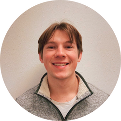 Close up portrait of tutor Nathan wearing a grey jacket and standing in front of a white wall.